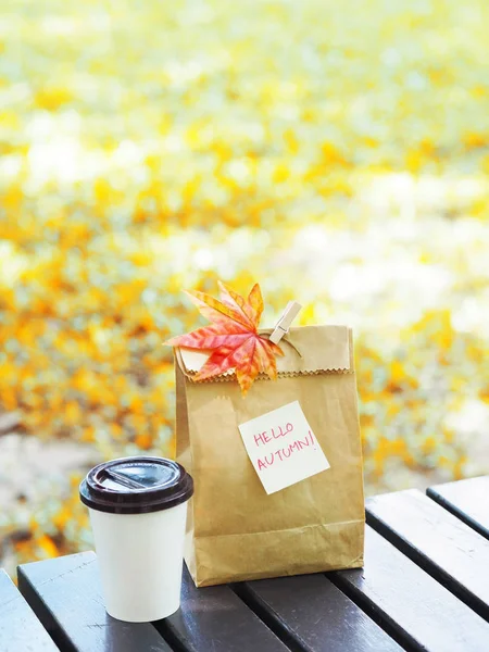 Take away breakfast in autumn. cup of hot coffee and brown paper bag with note and maple leaf on wooden table in the park.