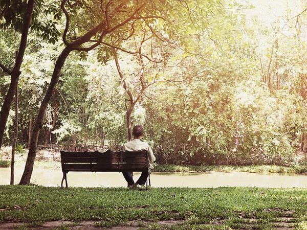 Back view of lonely old man siiting on wooden bench in the public park at sunrise. Vintage filter effect.