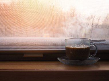 Yağmurlu bir sabahta cam pencerenin yanındaki ahşap masanın üzerine bir fincan kahve koy..