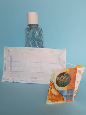 İsviçre banknotu 10 frank, yüz maskesi, jel alkollü şişe ve mavi arka plan.