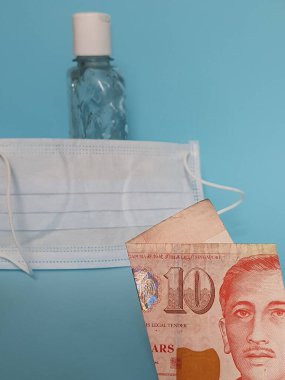 10 dolarlık Singapur banknotu, yüz maskesi, jel alkollü şişe ve mavi arka plan.