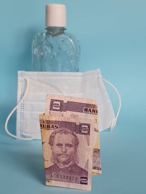 İki lempiralık Honduras banknotu, yüz maskesi, jel alkollü şişe ve mavi arka plan.
