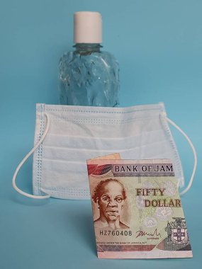 Jamaika 'da 50 dolarlık banknot, yüz maskesi, jel alkollü şişe ve mavi arka plan.