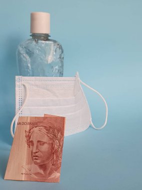 On reais 'lik Brezilya banknotu, yüz maskesi, jel alkollü şişe ve mavi arka plan.