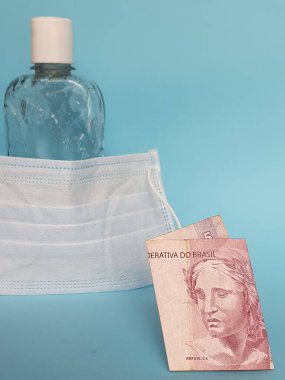 Brezilya banknotu beş reais, yüz maskesi, jel alkollü şişe ve mavi arka plan