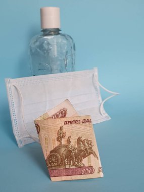 Rus banknotu 100 ruble, yüz maskesi, jel alkollü bir şişe ve mavi arka plan.
