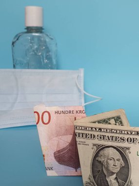 Bir dolarlık Amerikan banknotu, Norveç banknotu, yüz maskesi, jel alkollü bir şişe ve mavi arka plan.
