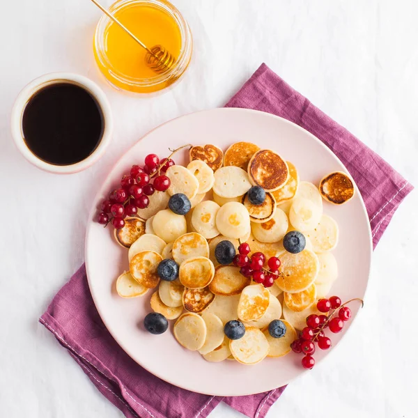 Tiny pancakes with berries, honey and coffee on a white tablecloth and space for text. Pancake cereal. The concept of Breakfast, food trends. Copy space. . High quality photo