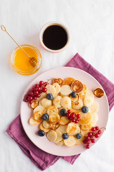 Tiny pancakes with berries, honey and coffee on a white tablecloth and space for text. Pancake cereal. The concept of Breakfast, food trends. Copy space. . High quality photo