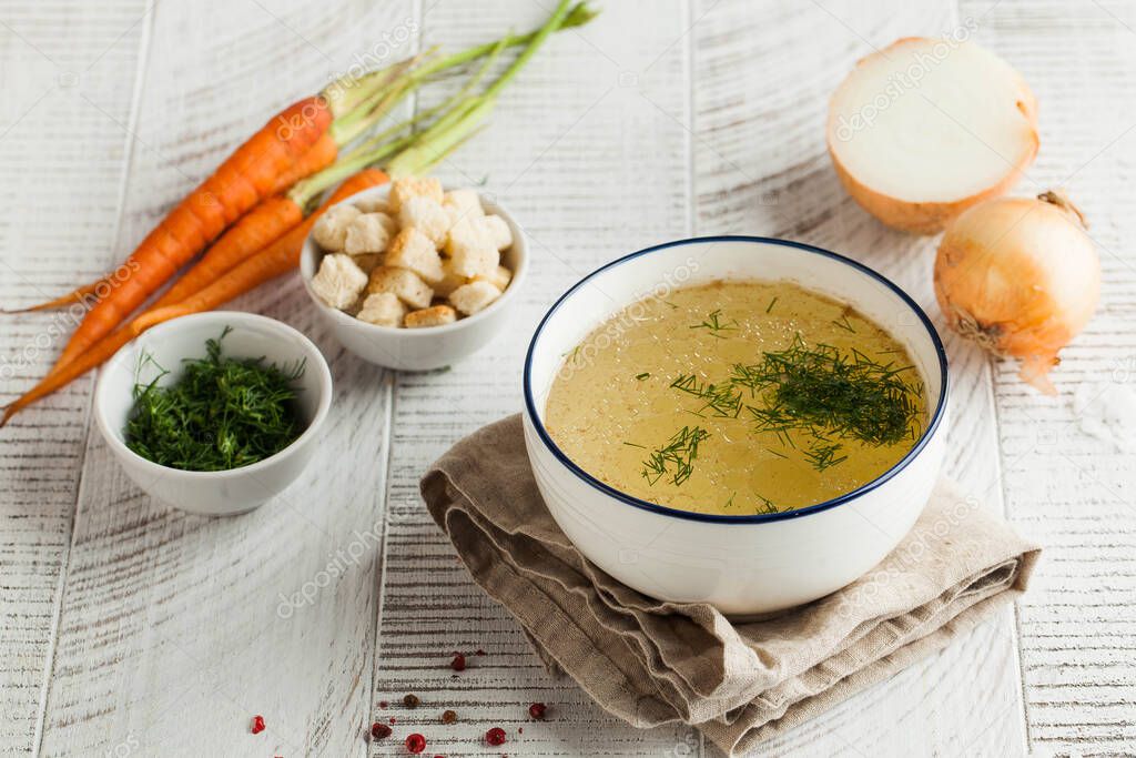 Homemade broth in a white bowl on a napkin with dill, fresh carrots and onions on a wooden background.