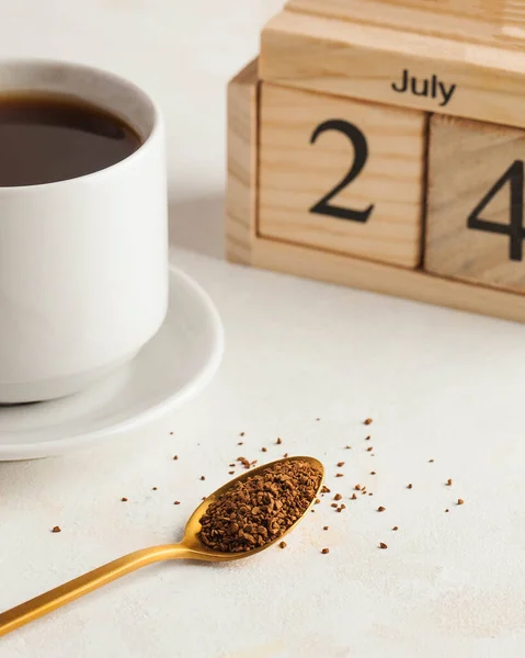 Instant coffee in a spoon on the background of a coffee mug and a wooden calendar on July 24. Birthday of instant coffee. High quality photo