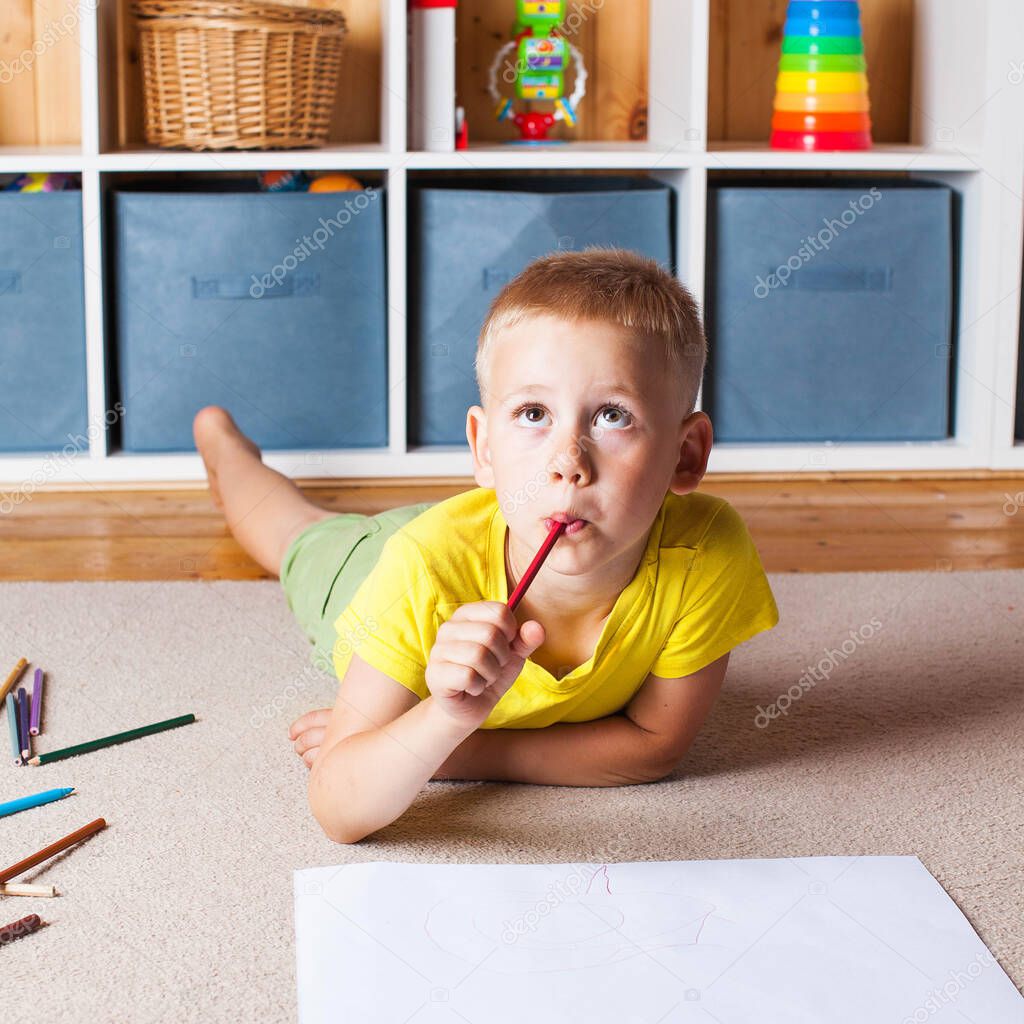 A cute blond boy in a yellow t shirt is lying on the floor with a sheet of paper and pencils and looks up thoughtfully. The concept of educational games, extracurricular learning.