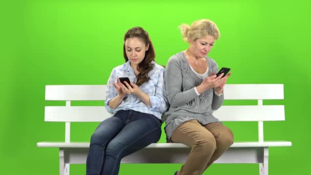 Una chica y una mujer están sentadas en un banco y mirando los teléfonos. Dependencia móvil. Pantalla verde — Vídeos de Stock