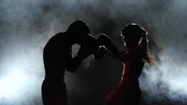 Um tipo com uma rapariga de capacetes e luvas de boxe a bater no ringue no escuro. Silhueta. Fundo de fumo. Movimento lento — Vídeo de Stock
