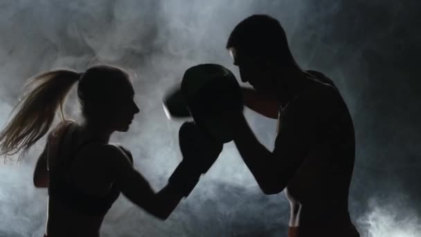 Un tipo con guantes de boxeo de una chica golpeando en el ring en la oscuridad. Silueta. Fondo de humo. Movimiento lento — Vídeo de stock