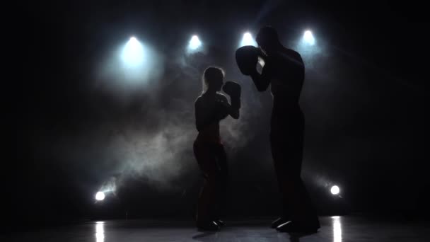 Tipo con una chica guantes de boxeo latiendo en el ring en la oscuridad, se están preparando para una competencia de kickboxing. Silueta. Luz por detrás. Fondo de humo. Movimiento lento — Vídeos de Stock