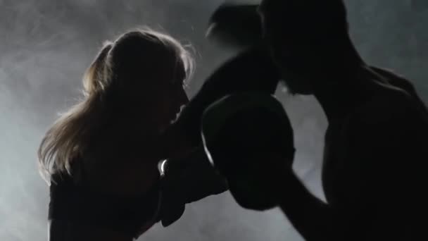 Un tipo con guantes de boxeo de una chica golpeando en el ring en la oscuridad. Silueta. Fondo de humo. Movimiento lento — Vídeo de stock