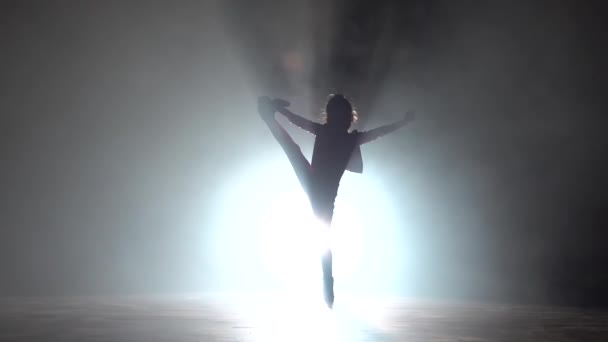 Bebé baila para sentarse en el cordel y levanta la pierna. Fondo de humo negro. Silueta. Luz por detrás. Movimiento lento — Vídeo de stock