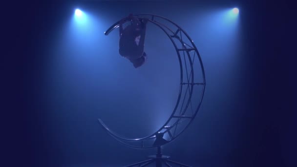 Гімнастка висить догори дном шпагатом на обертається металева конструкція місяця. Синій дим фону — стокове відео