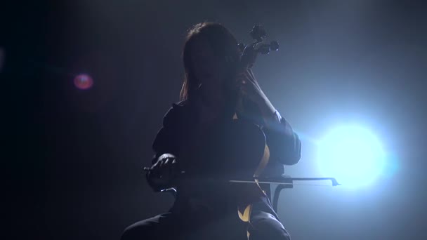 El músico en una habitación oscura se sienta en una silla y toca el violonchelo. Silueta. Fondo de humo negro — Vídeos de Stock