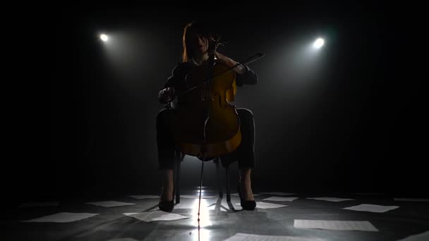Notas dispersas en el suelo y la silueta de un músico con un violonchelo en un estudio oscuro. Fondo de humo negro — Vídeo de stock