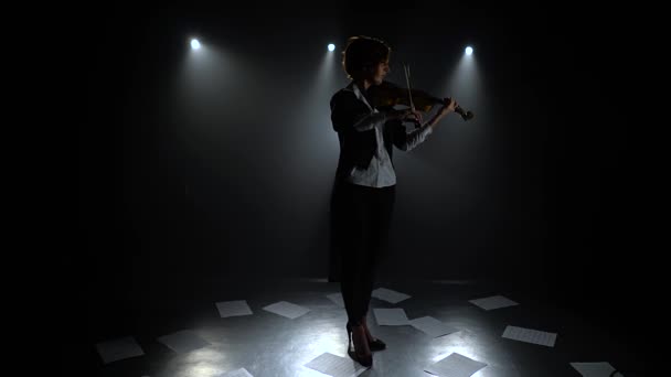 Chica del estudio oscuro tocando el violín en las hojas de notas. Silueta. Fondo de humo negro — Vídeo de stock