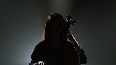 Müzisyen karanlık bir odada bir sandalyeye oturur ve viyolonsel çalıyor. Siluet. Siyah duman arka plan