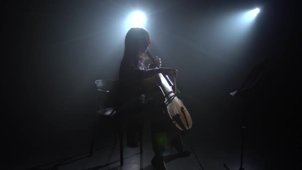 La mano femenina toca con un lazo en un violonchelo en un estudio oscuro. Silueta. Fondo de humo negro — Vídeos de Stock