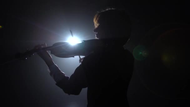 Profesional tocando el violín en un estudio oscuro con una linterna de él deslumbramiento. Fondo de humo negro. Vista trasera — Vídeos de Stock