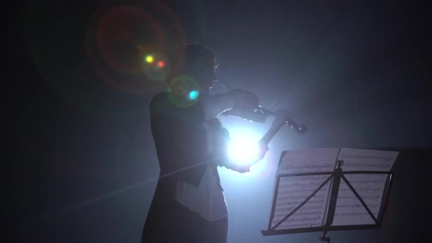 Девушка играет на скрипке и смотрит на стойку для музыки в темноте. Силуэт. Чёрный дым — стоковое видео