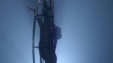 Acrobat bir metal yapının ayda dikey bir dize döndürür. Siyah duman arka plan. Ağır çekim. Yakın çekim