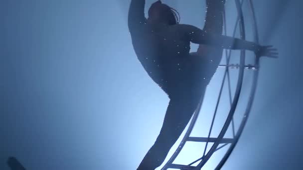 Girl in a brilliant costume performs the slips on one arm in the air on a metal rotating structure . Blue smoke background. Slow motion. Close up — Stock Video