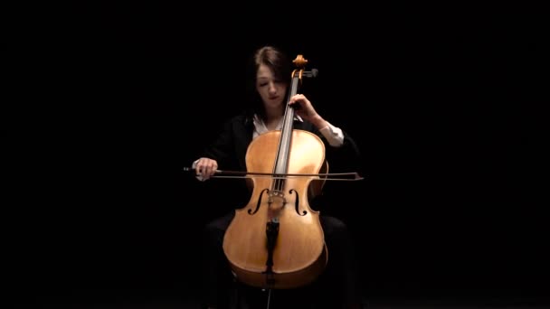 Dívka hraje na mistrovské violoncello sedě na židli v temné místnosti. Černé pozadí — Stock video