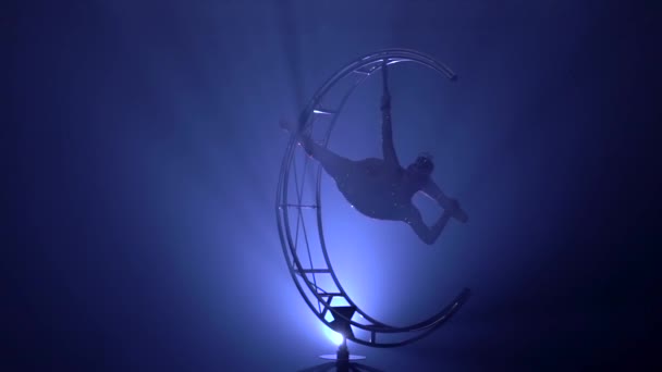 Gymnaste pendu à l'envers dans une ficelle sur une lune de structure tournante. Fond de fumée bleue. Mouvement lent.Silhouette 