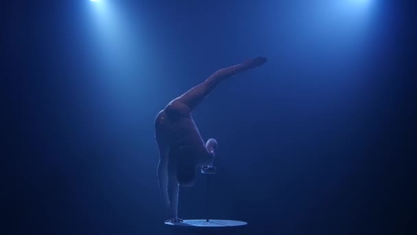 Chica gimnasta en la mesa muestra acrobacias. Fondo azul humo. Movimiento lento — Vídeo de stock