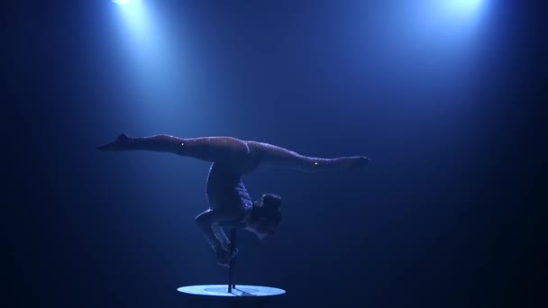 Acrobat girl on stage does acrobatic stunts standing on her hands on the table. Smoke blue background. Slow motion. Silhouette — Stock Video