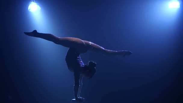 Acrobat meisje op het podium doet acrobatische stunts permanent op handen. Roken blauwe achtergrond. Slow-motion. Silhouet — Stockvideo