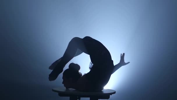 Akrobatin dreht sich auf einem Tisch, der auf dem Kopf steht. Rauchentwicklung. Zeitlupe. Silhouette — Stockvideo