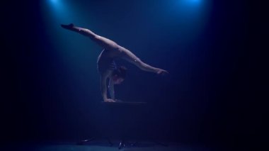 Jimnastikçi akrobasi karanlık bir stüdyoda bir tablo üzerinde gerçekleştirir. Mavi renkli duman. Ağır çekim