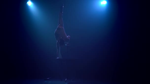 Acrobat ragazza sul palco fa acrobazie acrobatiche in piedi sulle sue mani sul tavolo. Fondo blu fumo. Al rallentatore. Silhouette — Video Stock