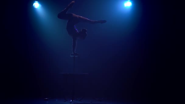 Девушка-акробатка на сцене делает акробатические трюки, стоя на руках на столе. Дым на голубом фоне. Медленное движение. Силуэт — стоковое видео