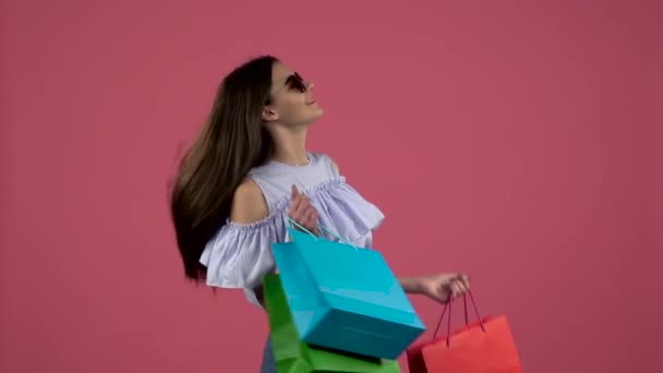 Gir em óculos de sol l depois de fazer compras dançando com pacotes coloridos. Fundo rosa. Movimento lento — Vídeo de Stock