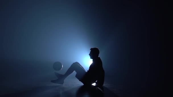 Fotboll freestyle. Freestyler göra tricks sitter på golvet mot blå Spotlight. Slow motion — Stockvideo