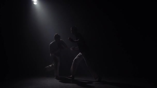 スタジオでスポットライトに対して暗闇の中でカポエイラを練習するアスレチックの男たち. — ストック動画