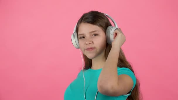 Adolescente mujer escucha música y baila en grandes auriculares blancos sobre fondo rosa. Movimiento lento — Vídeo de stock