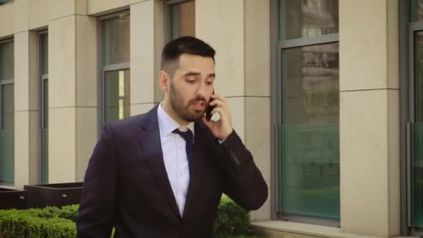 Giovane uomo d'affari si sente infastidito mentre parla al telefono cellulare all'aperto vicino al centro dell'edificio per uffici. Uomo arrabbiato e triste . — Video Stock