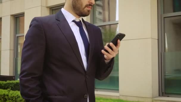 Vue rapprochée d'un homme d'affaires souriant vêtu d'une chemise à l'aide d'un smartphone alors qu'il se promène sur le fond d'un bâtiment moderne — Video