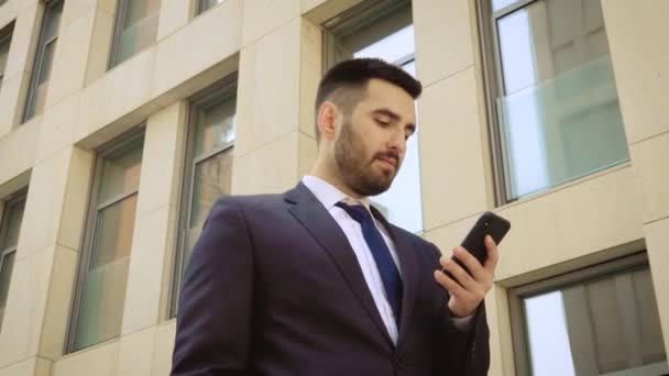 Vista de cerca de un hombre de negocios sonriente vestido con camisa usando un teléfono inteligente mientras camina sobre el fondo de un edificio moderno — Vídeos de Stock