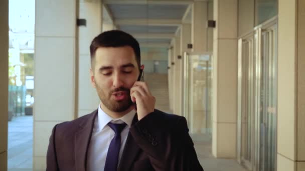 Vista de cerca del hombre de negocios vestido con camisa hablando smartphone mientras camina sobre el fondo de un edificio moderno — Vídeos de Stock