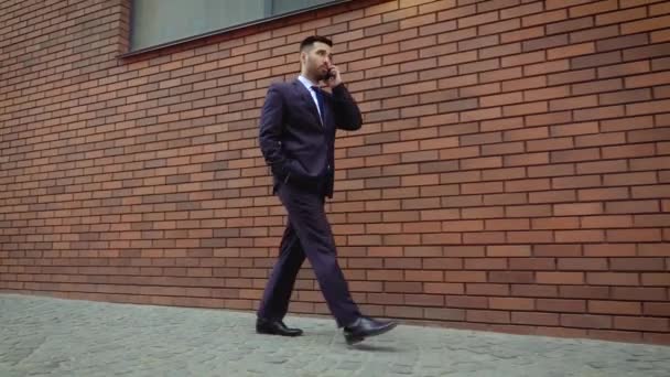 Young businessman feeling annoyed while talking on the cell phone outdoors near modern brick wall. Man angry and sad. — Stock Video